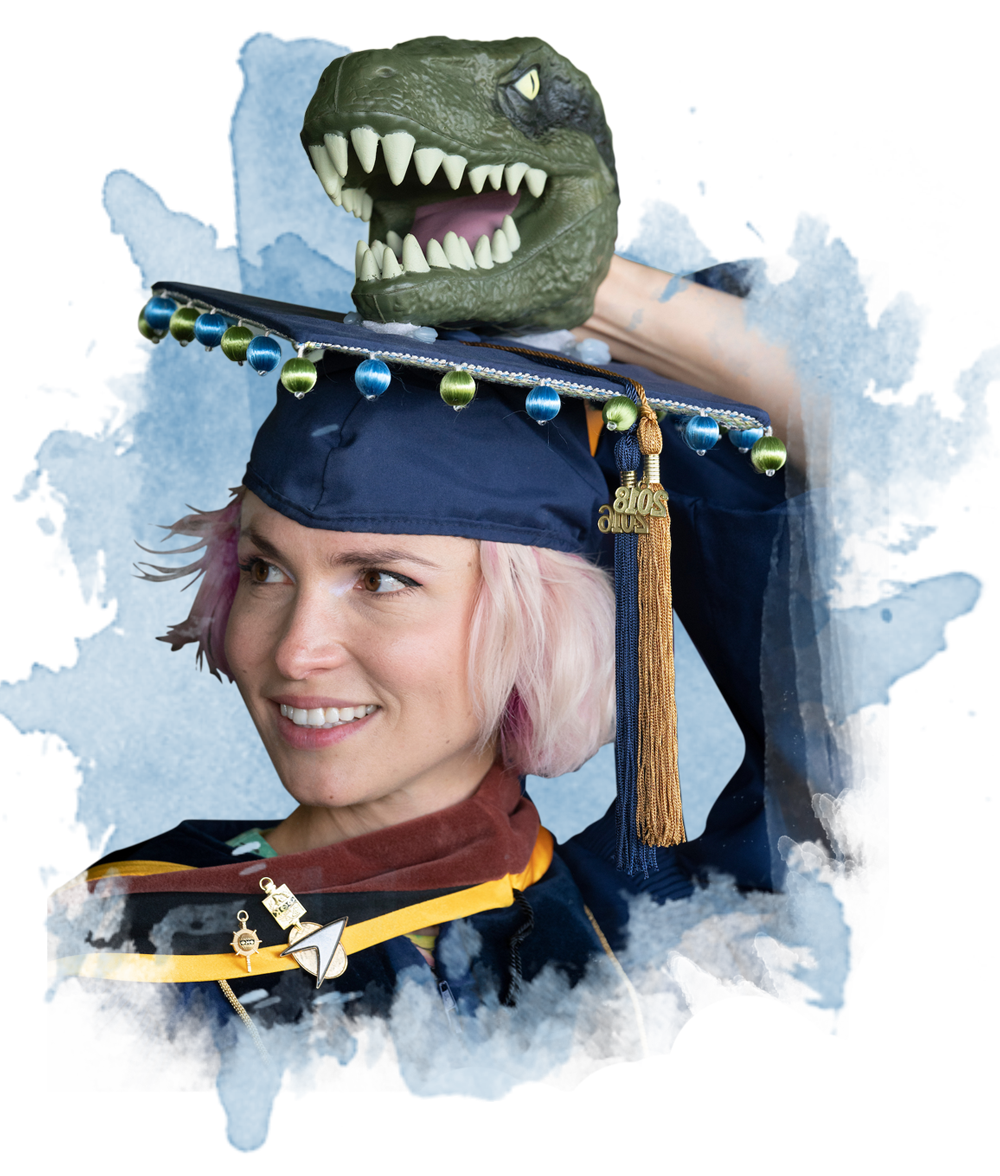 A student dressed for graduation with a t-rex head atop her grad cap