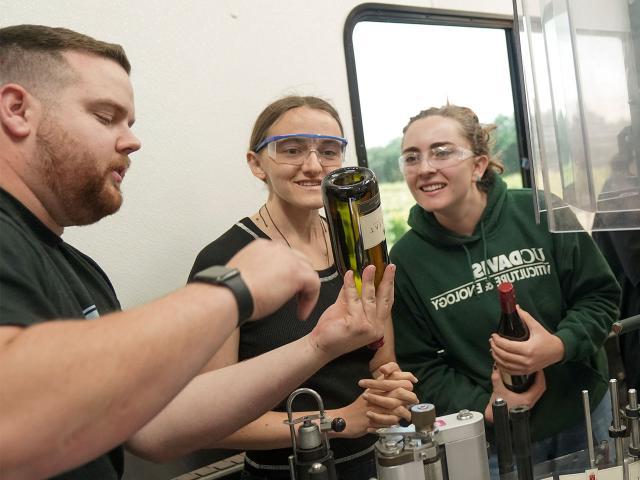 A Halsey Bottling employee holds up a wine bottle and points to it while explaining a point to two smiling students in safety goggles.