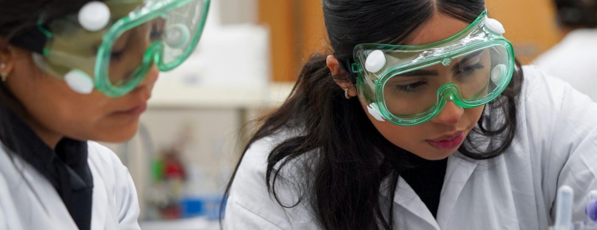 在amjs澳金沙门，两名化学专业的学生在实验室工作台上戴着护目镜处理药物