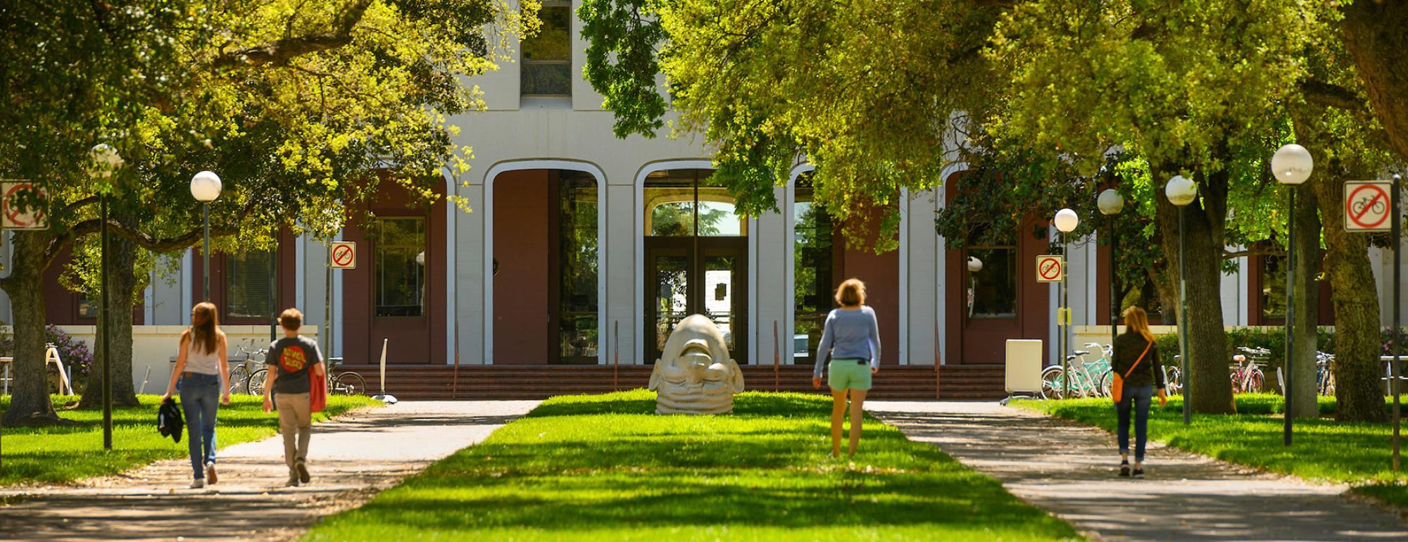 Four people walking in front of Mrak Hall on campus. One of the Egghead statues 'Eye on Mrak' is in the background.