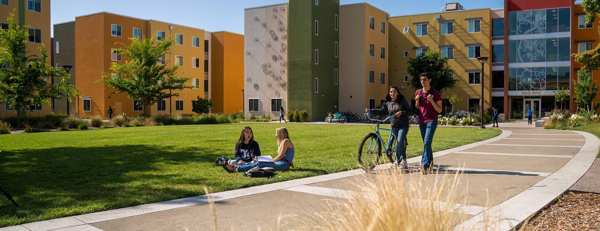 two student walking a bike while two more students are sitting on the grass in front of the residence halls