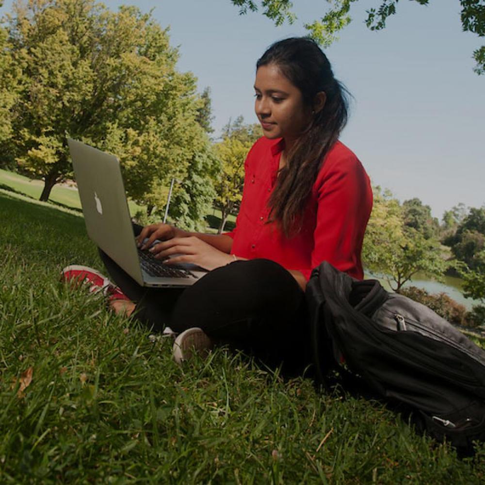 A female student in red reads from her laptop on the UC Davis quad