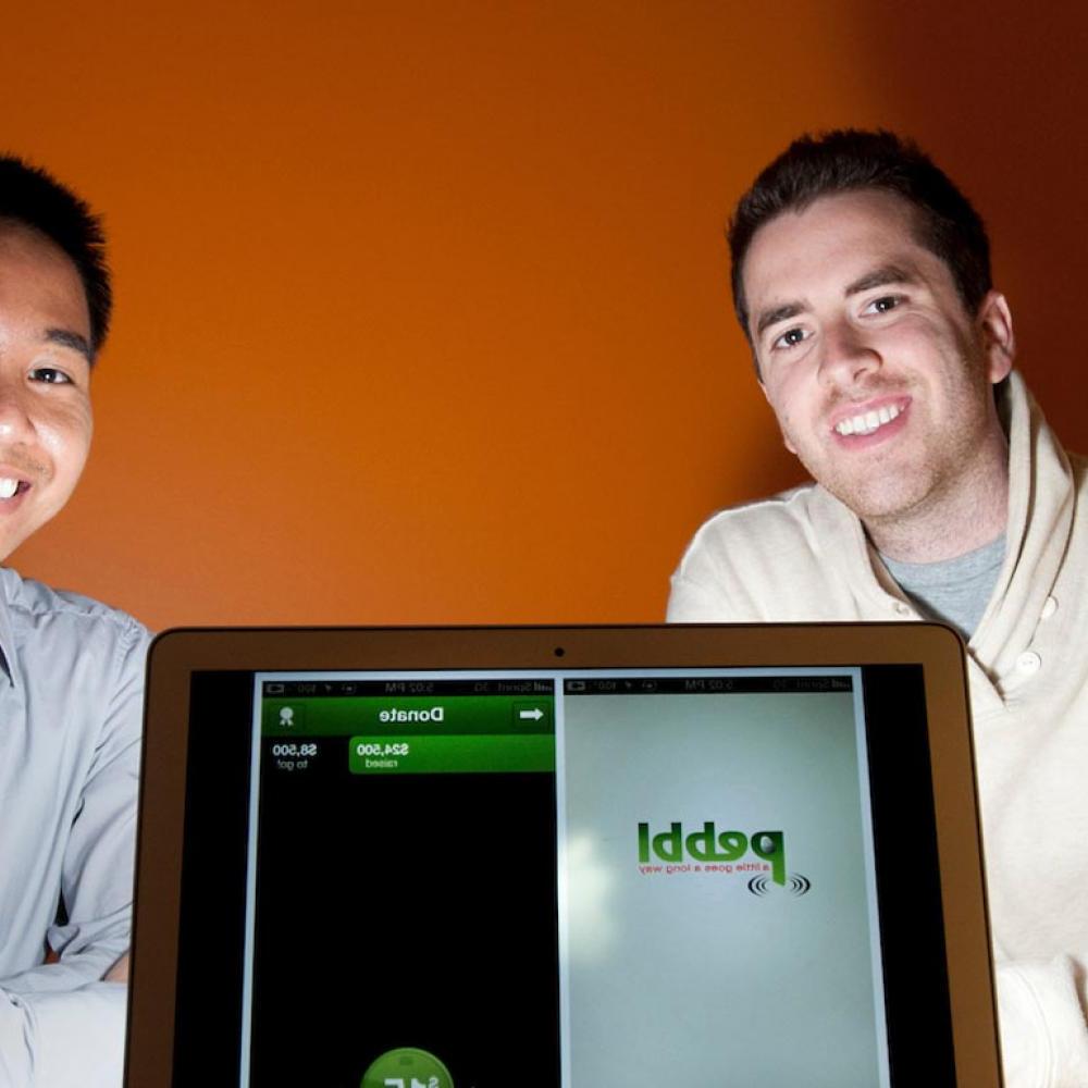 两个研究生拿着一台笔记本电脑摆姿势，上面显示着他们开发的应用程序.