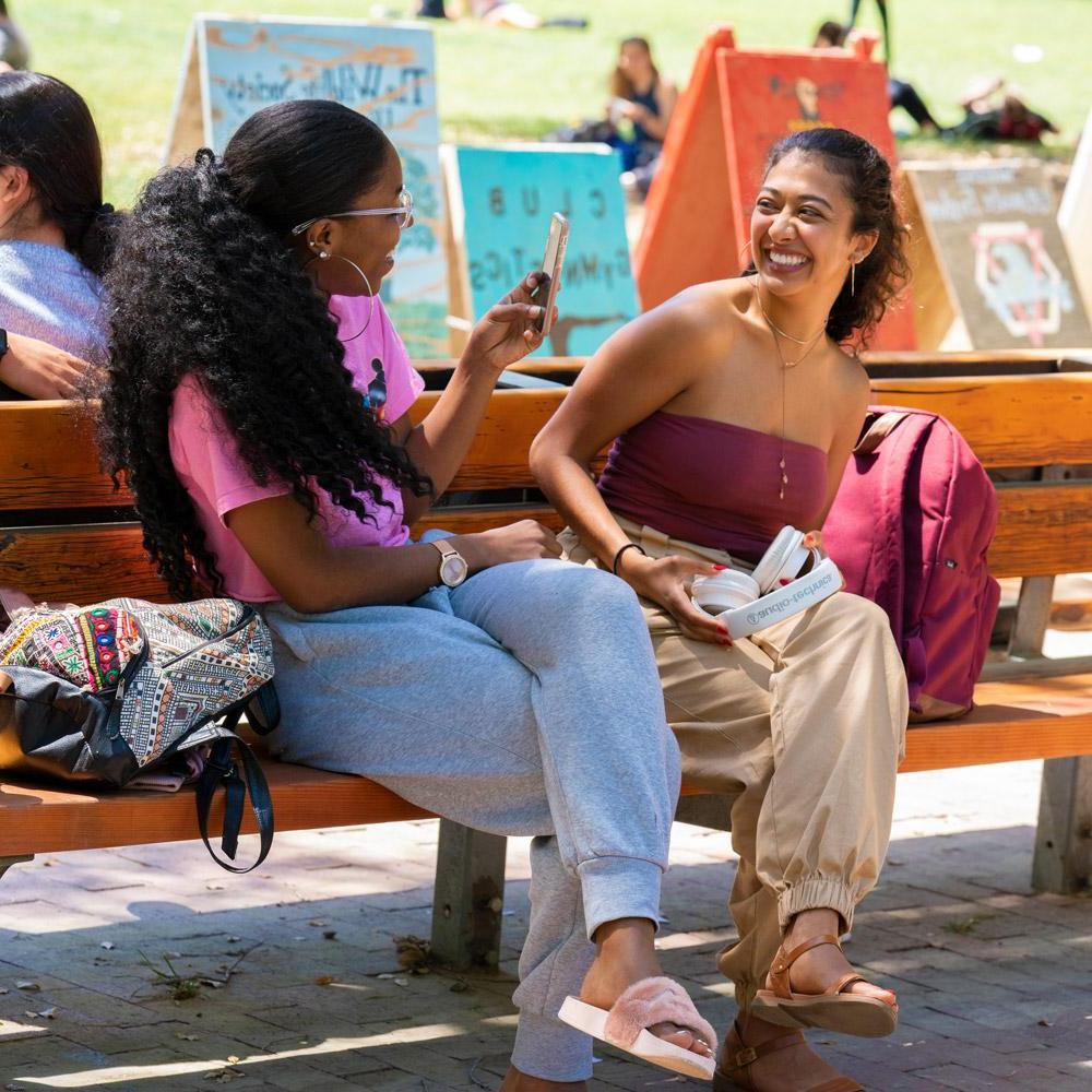 two students sitting on a bench near the quad with signs of student organizations in the background
